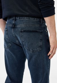 Vaqueros de mezclilla oscura con corte ajustado, bolsillos traseros visibles y diseño de pierna recta. Presenta un ligero desgastado y una cinturilla clásica.