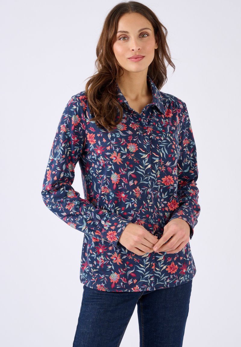 Donna con lunghi capelli castani che indossa una camicia a maniche lunghe blu navy con bottoni, decorata con motivi floreali rossi e rosa, abbinata a jeans blu scuro.