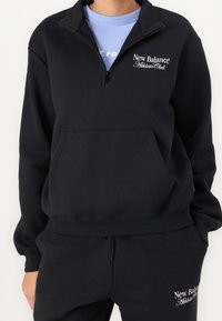 Zwarte sweatvest met ritssluiting en hoge kraag, voorzien van een voorvak en het logo "New Balance Athletic Club" in wit. Zachte stoftextuur.