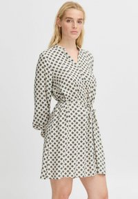 Rochie cu butoane, cu model, cu mâneci lungi, având un fundal crem și modele spirale negre. Detaliul cu talie legată evidențiază silueta.