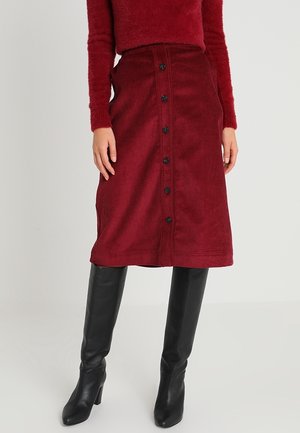 Kvinde iført en rød manchetknap-midi nederdel i fløjl, rød, blød langærmet top og sorte knæhøje støvler med hæl.