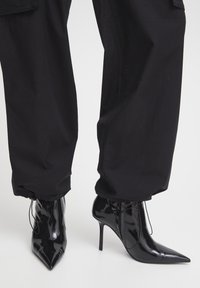 Čierne lakované kotníkové čižmy s špičatými prstami a vysokými stiletto podpätkami, kombinované s voľnými čiernymi nohavicami.