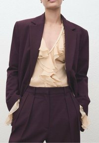 Veste de costume bordeaux sur un chemisier beige à volants, agrémentée d'un collier de perles. Pantalon tailleur à plis, textures contrastées sur l'ensemble.