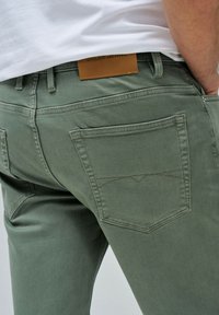Pantalones de mezclilla verde de algodón con corte recto, que presentan un bolsillo trasero con detalles de costura y una etiqueta de marca de cuero visible.