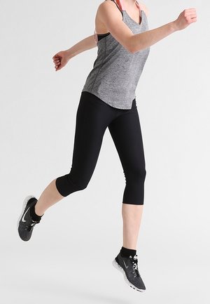 Nainen harmaassa liivipaidassa ja mustissa legginsseissä kesken hyppyyn, päällään mustat Nike-juoksukenkiä, yksinkertaista valkoista taustaa vasten.