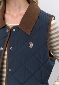 Quiltet vest i marineblå med brun kordfløyelkrage. Har trykknapper og en liten brodert logo på forsiden. Stripete skjorte er synlig under.