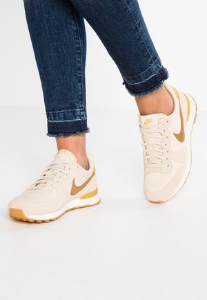 Personne portant des baskets Nike beige et moutarde associées à un jean bleu foncé à ourlet effiloché sur un sol blanc.
