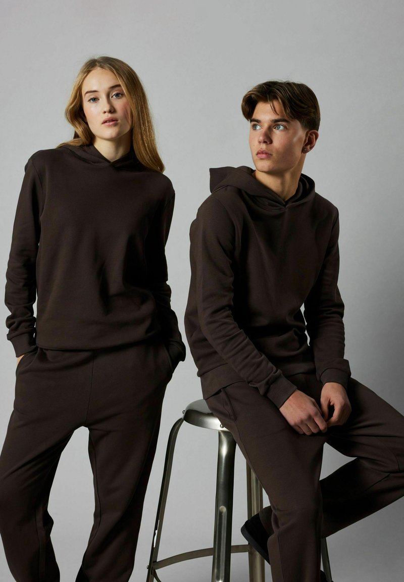 Brūnas kapuces sporta džemperis un bikses, kas izgatavotas no mīksta materiāla; divi modeļi pozē, viens sēž uz metāla krēsla, neitrāls pelēks fons.