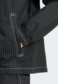 Ruka v boční kapse černého pruhovaného saka s bílým prošív áním a elastickým manžetovým zakončením na rukávu.