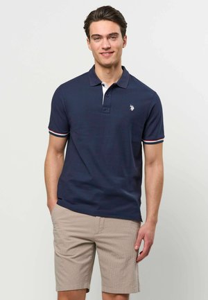 Fiatal férfi tengerészkék pólóingben, fehér logóval és bézs rövidnadrágban, egyszerű, világos háttér előtt áll, enyhén mosolyogva.