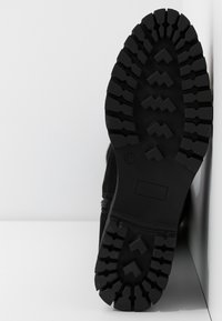 Semelle de botte noire avec un motif de relief profond et robuste reposant contre un coin blanc de mur et de sol.
