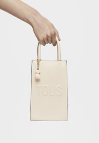 Bolso tote beige con el logo "TOUS" en relieve, textura suave, asas redondeadas en la parte superior y un charm de oso colgante en rosa claro.