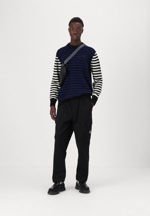 Sweter w paski w ciemnoniebieskie i czarne linie, w połączeniu z czarnymi spodniami cargo. Czarna torba zawieszona na ramieniu. Czarne buty dopełniają całość.