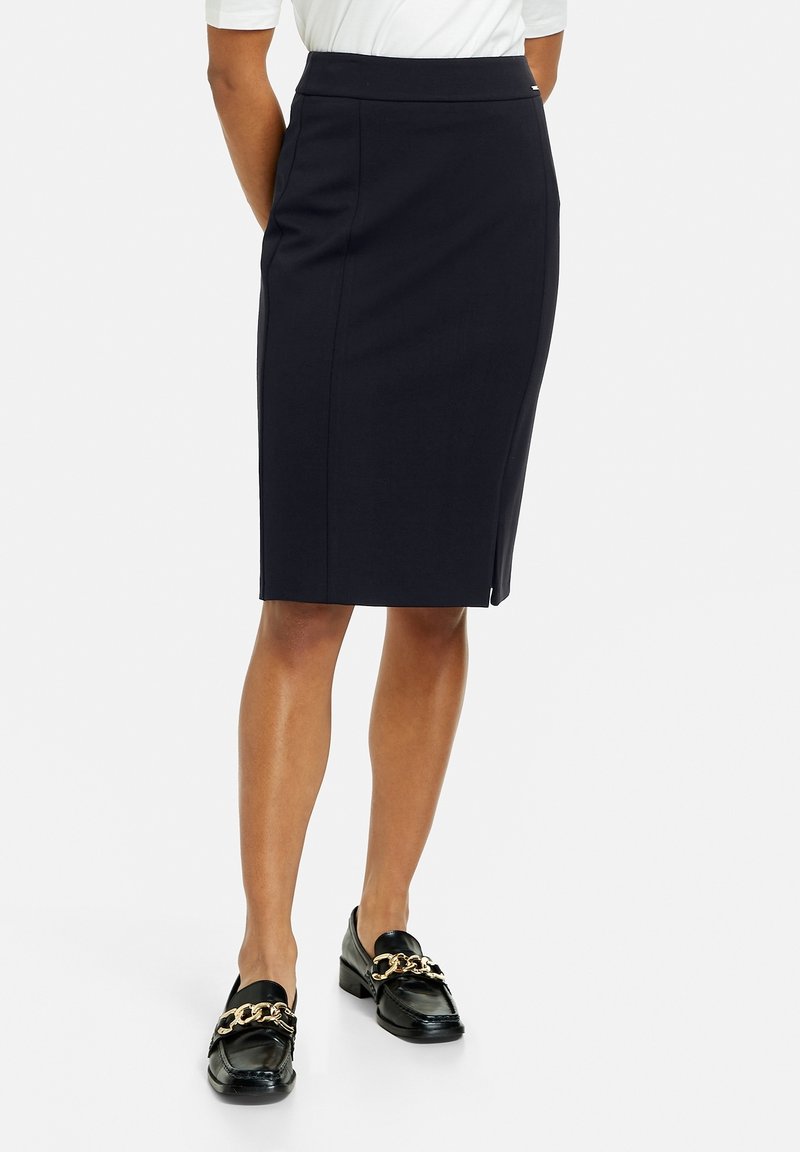Femme portant une jupe crayon bleu marine longueur genou, un haut blanc et des mocassins noirs avec des détails en chaîne dorée, debout sur un fond uni.