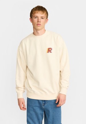 Jeune homme portant un sweat crème avec un « R » multicolore sur la poitrine et un jean bleu, debout devant un fond uni.