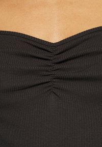 Pelle nuda sopra un top in tessuto nero a coste con pieghe verticali lungo il centro del colletto anteriore.