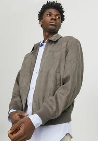 Jachetă bomber din suede gri, cu manșete și guler ribbed; are un închidere prin fermoar și două buzunare laterale, purtată peste o cămașă de culoare albastru deschis.