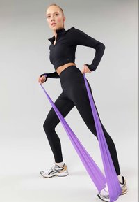 Junge Frau in schwarzer Sportbekleidung, die ein lila Widerstandsband unter dem Fuß dehnt, während sie in einem weiten Ausfallschritt steht, vor einem schlichten Hintergrund.