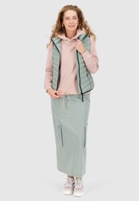 Gilet verde imbottito sopra un felpa rosa, abbinato a una maxi gonna verde chiaro con tasche con zip e scarpe da ginnastica leggere.