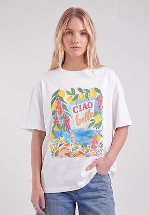 Donna bionda che indossa una maglietta bianca con stampa colorata di limoni, fiori, edifici e testo "CIAO bella", in piedi con una mano nella tasca dei jeans blu.