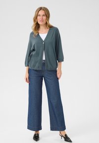 Cardigan vert forêt avec un col en V, des manches trois-quarts et une fermeture à un bouton, associé à un pantalon en denim à jambes larges.