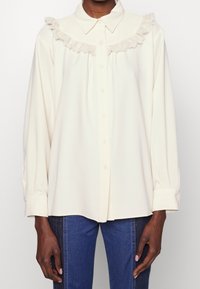 Cream button-up shirt with a ruffled lace collar, long sleeves, and a relaxed fit. Paired with blue denim jeans.