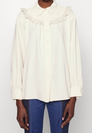 Camisa crema de botones con cuello de encaje con volantes, mangas largas y un corte relajado. Combinada con jeans de mezclilla azul.