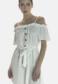 Vestido blanco sin hombros con cuello con volantes, tres botones marrones, cintura elástica y cinta de tela. Textura suave y corte a la altura de la rodilla.