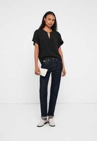 Woman wearing black short-sleeve top, dark rolled-up jeans, white slip-on shoes, holding a small white textured clutch, standing against a plain background.