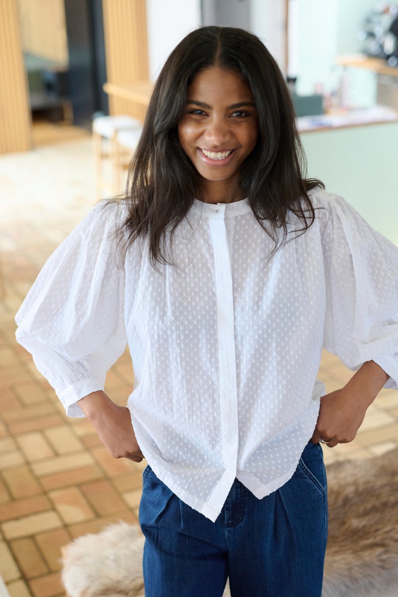 Jeune femme souriante, portant un chemisier blanc texturé à manches longues et un pantalon bleu, debout à l'intérieur avec les mains sur les hanches.