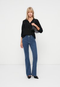 Black open-knit blouse with a V-neck. Blue denim flared jeans with white vertical stripes. Model holding a black handbag and wearing black shoes.