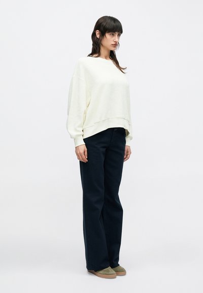 Ανοιχτό κρεμ φαρδύ φούτερ με ριμπ μανσέτες, σε συνδυασμό με σκούρο μπλε παντελόνι με φαρδιά πόδια και πράσινα σουέντ παπούτσια. Απλή, χαλαρή εφαρμογή.