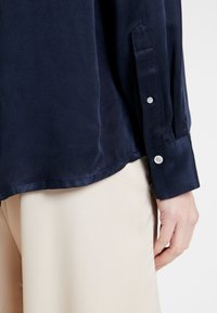 Camicia leggera blu navy a maniche lunghe con polsini a bottoni e una texture liscia, abbinata a pantaloni beige.