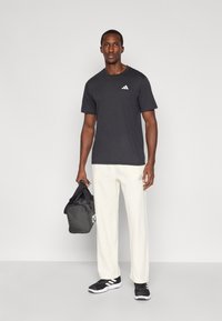 Roupa atlética composta por uma camisola preta de mangas curtas com um logótipo branco, calças brancas texturizadas e sapatilhas pretas de desporto. A portar uma mala de ginásio preta.