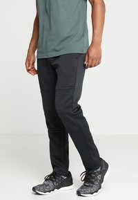 Svarta träningsbyxor med ribbade sidopaneler, tillsammans med en grön t-shirt. Tyget verkar slätt med en figurnära design. Svarta träningsskor.