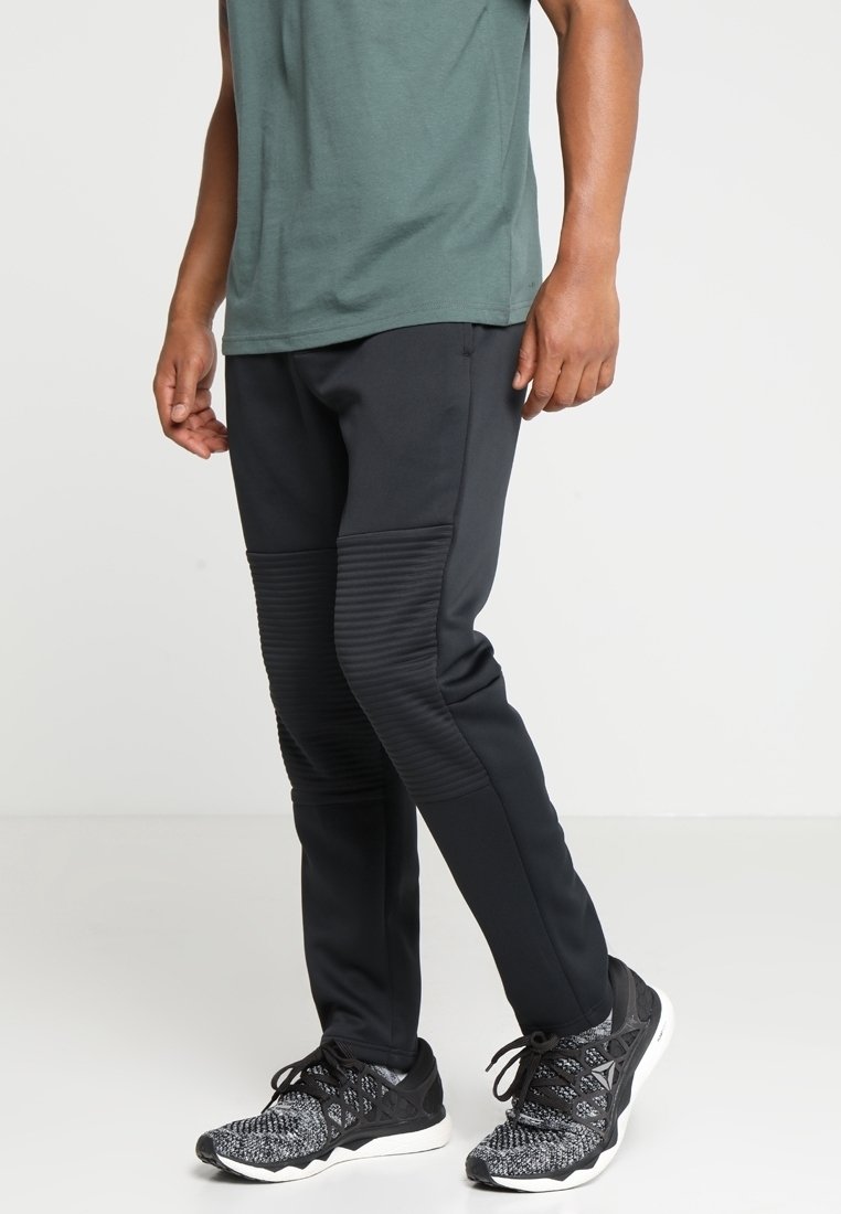 Svarta träningsbyxor med ribbade sidopaneler, tillsammans med en grön t-shirt. Tyget verkar slätt med en figurnära design. Svarta träningsskor.