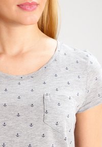 Camiseta gris de manga corta hecha de una tela suave con escote redondeado, que presenta un patrón repetido de anclas en azul marino y un pequeño bolsillo en el pecho.
