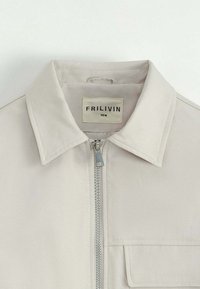 Veste beige clair avec un col, fermeture éclair frontale argentée, et une poche à rabat sur le côté droit de la poitrine.