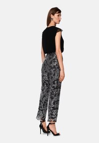 Woman in black sleeveless top, silver sequin wide-leg pants, black high-heeled sandals, and silver earrings, standing against white background.