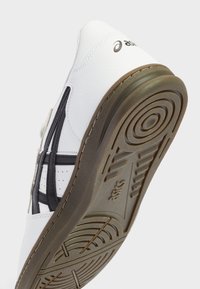 Chaussure de sport blanche avec une tige en cuir texturé, des accents noirs et une semelle en caoutchouc présentant un motif en spirale. Détails perforés uniques visibles.
