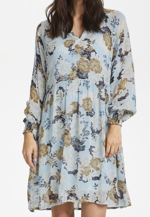 Femme portant une robe bleu clair à manches longues et longueur genoux, avec des motifs floraux beige et bleu foncé et des poignets froncés.