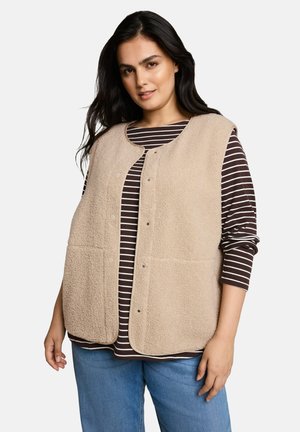 Frau trägt eine beige Fleeceweste über einem braun-weiß gestreiften Langarmshirt und blauen Jeans, steht vor einem einfarbigen Hintergrund.