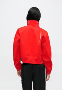 Rdeča skrajšana jakna z visokim ovratnikom, narejena iz sijočega materiala. Ima elastične manšete in spodnji rob, gladko teksturo in elegantno zasnovo.