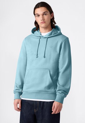 Junger Mann mit schulterlangem Haar, der einen hellblauen Hoodie und dunkle Jeans trägt und vor einem einfarbigen weißen Hintergrund steht.