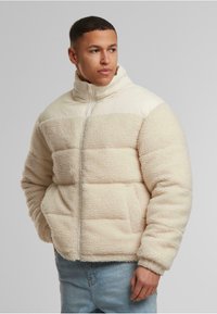 Beige dunjakke med en teksturert ytterlag og glatt øverste seksjon. Har høy krage, glidelåslukking og sidelommer.