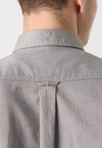 Grijze stoffen shirt met een button-down kraag, met een rugplooi en gestikte details. De textuur lijkt glad en gelijkmatig.