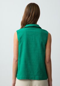 Blusa verde senza maniche con colletto, realizzata in un tessuto testurizzato e con fessure laterali all'orlo, vista da dietro.
