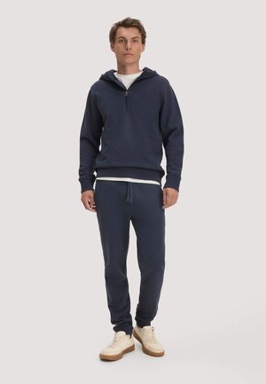 Junger Mann steht, trägt einen marineblauen Hoodie mit Frontreißverschluss, passende marineblaue Jogginghosen, weiße Sneaker und ein weißes Shirt darunter.