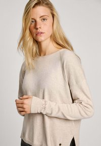 Beige neulepaita, jossa on leveä kaula-aukko, pitkät hihat ja sivuhalkiot. Sisältää brodeerattua yksityiskohtaa hihoissa ja pienen logoesitteen.