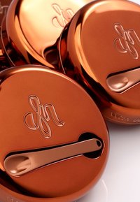 Kuparinvärinen kompatti, jossa on tasainen, heijastava pinta. Ylhäällä on pieni lasta ja tyylikäs logo kohokuviolla.